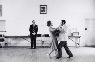 David Goldblatt; Dancing-master Ted van Rensburg watches two of his ballroom pupils, swinging to the music of a recording of Victor Sylvester and his Orchestra, in the hall of the Memorable Order of Tin Hats, at the old Court House, Boksburg, Transvaal. May 1980. Image size: 31 by 46cm.